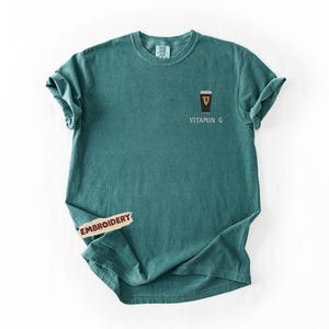 May include: Teal-colored t-shirt with a small embroidered design of a pint of stout beer and the words "Vitamin G". The shirt has short sleeves and a crew neck. The shirt is made of a soft, comfortable material.