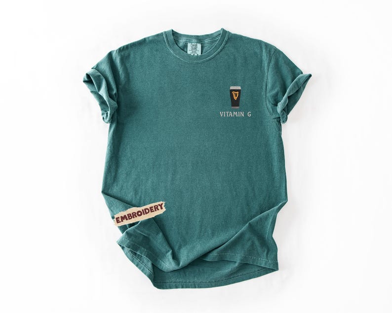 May include: Teal-colored t-shirt with a small embroidered design of a pint of stout beer and the words "Vitamin G". The shirt has short sleeves and a crew neck. The shirt is made of a soft, comfortable material.