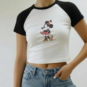 Puede incluir: Top corto blanco con mangas ragl&aacute;n negras con un gr&aacute;fico de Minnie Mouse. El personaje de dibujos animados lleva un vestido rojo con lunares blancos y zapatos marrones. La camiseta se combina con unos vaqueros azules.