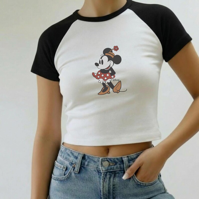 Puede incluir: Top corto blanco con mangas ragl&aacute;n negras con un gr&aacute;fico de Minnie Mouse. El personaje de dibujos animados lleva un vestido rojo con lunares blancos y zapatos marrones. La camiseta se combina con unos vaqueros azules.