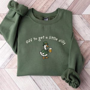 May include: Green sweatshirt with the text "Off to get a little silly." A cartoon duck wearing a green hat and jacket is holding a beer mug. The sweatshirt is a solid green color.