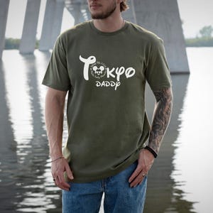 May include: Olive green t-shirt with white text that reads "Tokyo Daddy" in a stylized font. The shirt features a Mickey Mouse graphic and is paired with blue jeans. The person is standing in front of a bridge and water.