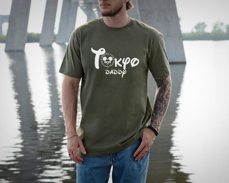 May include: Olive green t-shirt with white text that reads "Tokyo Daddy" in a stylized font. The shirt features a Mickey Mouse graphic and is paired with blue jeans. The person is standing in front of a bridge and water.