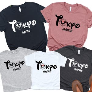 May include: Five t-shirts in various colors: navy, dusty rose, gray, white, and charcoal. Each shirt features the word "Tokyo" in a stylized font, with a Mickey Mouse graphic and the word "Name" below. Sandals are at the bottom.