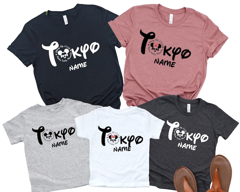 May include: Five t-shirts in various colors: navy, dusty rose, gray, white, and charcoal. Each shirt features the word "Tokyo" in a stylized font, with a Mickey Mouse graphic and the word "Name" below. Sandals are at the bottom.