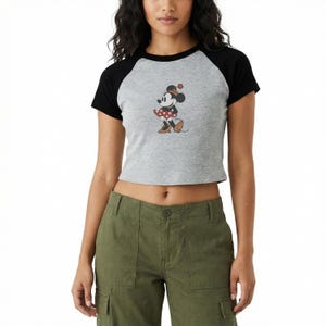 Puede incluir: Top corto gris ragl&aacute;n con mangas negras con un gr&aacute;fico de Minnie Mouse. El personaje de dibujos animados lleva un vestido de lunares rojos y blancos y un sombrero a juego. La camiseta se combina con pantalones cargo verde oliva.