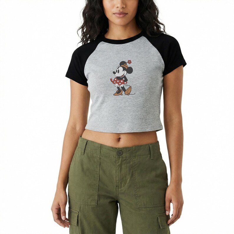 Puede incluir: Top corto gris ragl&aacute;n con mangas negras con un gr&aacute;fico de Minnie Mouse. El personaje de dibujos animados lleva un vestido de lunares rojos y blancos y un sombrero a juego. La camiseta se combina con pantalones cargo verde oliva.