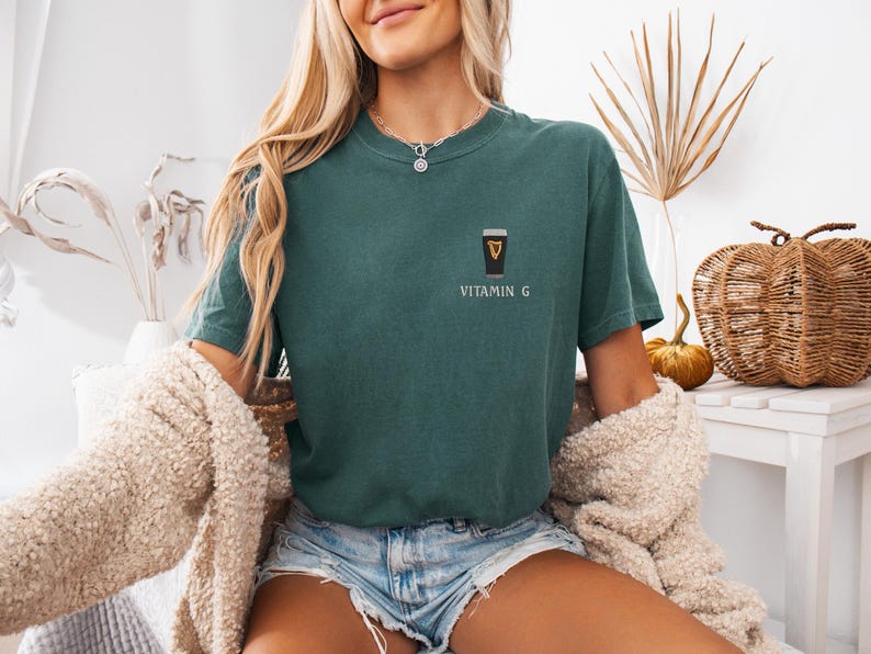 May include: Teal-colored t-shirt with a small graphic of a pint of stout and the text "VITAMIN G". The shirt is a short-sleeved, crew neck style. The model is wearing denim shorts and a cream-colored cardigan.