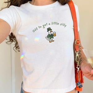 May include: White t-shirt with the phrase "Off to get a little silly" in green lettering. The shirt features a cartoon goose wearing a green hat and jacket, holding a beer mug. The shirt is short-sleeved.