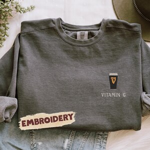 May include: Dark gray sweatshirt with a black and gold graphic of a pint of stout and the words "Vitamin G". The word "Embroidery" is printed on a tan banner. The sweatshirt is folded on top of blue jeans.