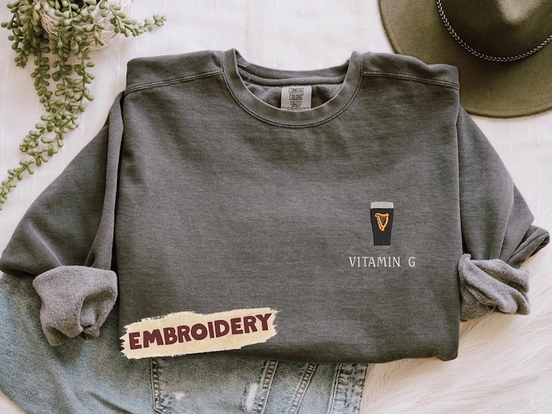 May include: Dark gray sweatshirt with a black and gold graphic of a pint of stout and the words "Vitamin G". The word "Embroidery" is printed on a tan banner. The sweatshirt is folded on top of blue jeans.