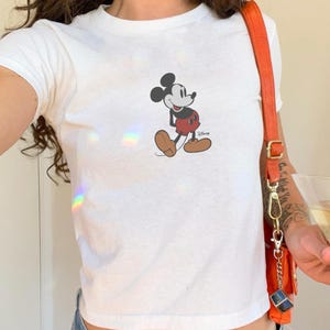 May include: White t-shirt featuring a classic Mickey Mouse graphic. The cartoon character is depicted in his iconic pose, wearing red shorts and brown shoes. The Disney logo is visible below the character.