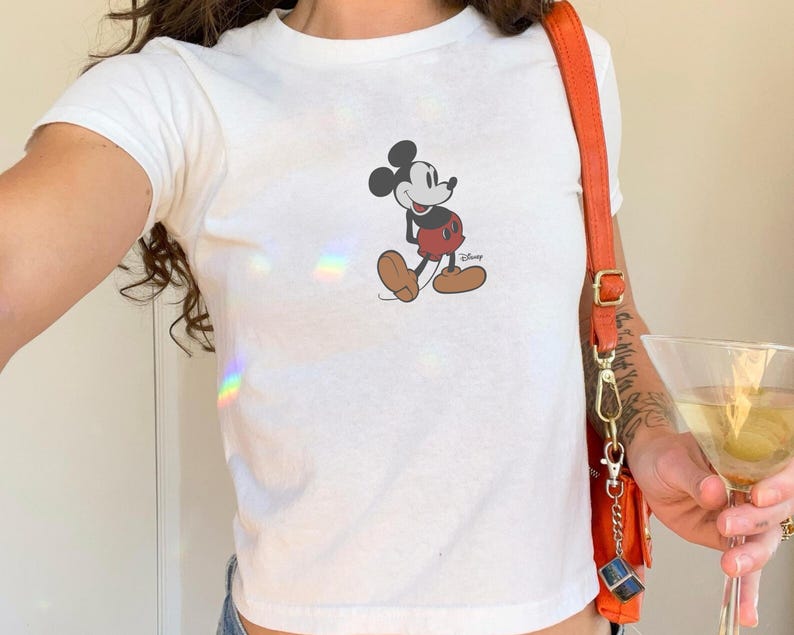 May include: White t-shirt featuring a classic Mickey Mouse graphic. The cartoon character is depicted in his iconic pose, wearing red shorts and brown shoes. The Disney logo is visible below the character.