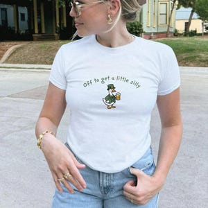 May include: White t-shirt with the phrase "Off to get a little silly" in green script. A cartoon goose wearing a hat and holding a beer mug is printed below the text. The shirt is short-sleeved.