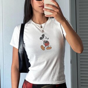 May include: White short-sleeved t-shirt featuring a classic Mickey Mouse graphic. The cartoon character is depicted in his iconic pose, wearing red shorts, yellow shoes, and white gloves. The shirt is a casual style.