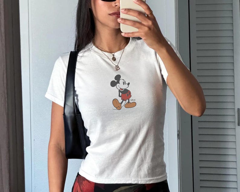 May include: White short-sleeved t-shirt featuring a classic Mickey Mouse graphic. The cartoon character is depicted in his iconic pose, wearing red shorts, yellow shoes, and white gloves. The shirt is a casual style.