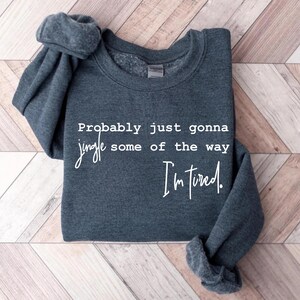 Puede incluir: Sudadera gris oscuro con el texto blanco: "Probably just gonna jingle some of the way I'm tired". La prenda est&aacute; doblada, con las mangas visibles. Sobre fondo de madera.