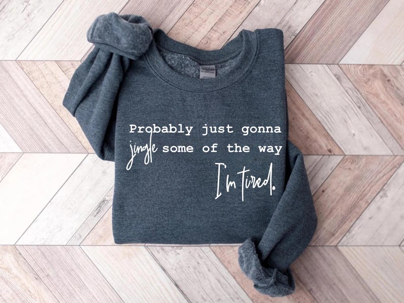 Puede incluir: Sudadera gris oscuro con el texto blanco: "Probably just gonna jingle some of the way I'm tired". La prenda est&aacute; doblada, con las mangas visibles. Sobre fondo de madera.