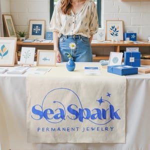 May include: A display table with a white tablecloth and a beige runner featuring the blue text "Sea Spark PERMANENT JEWELRY." The table holds framed art, blue gift boxes, and a small blue vase with a daisy. A person stands behind the table.