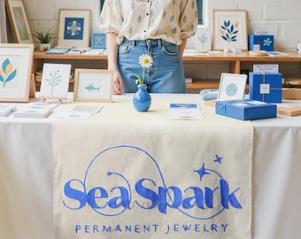 May include: A display table with a white tablecloth and a beige runner featuring the blue text "Sea Spark PERMANENT JEWELRY." The table holds framed art, blue gift boxes, and a small blue vase with a daisy. A person stands behind the table.