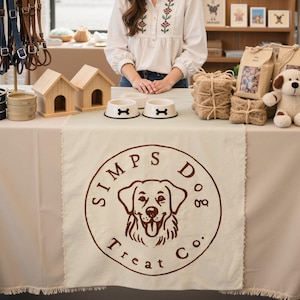 May include: A display of dog products, including bowls, leashes, and treats. The table features a beige tablecloth with the logo "Simps Dog Treat Co." The products include wooden dog houses, dog bowls with bone designs, and a stuffed dog toy.