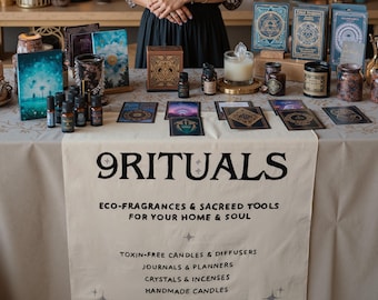 May include: A table display of spiritual and home decor items, including candles, essential oils, journals, and crystals. The table features a banner with the text "9 RITUALS" and product descriptions. Various products are arranged on a cream-coloured tablecloth.