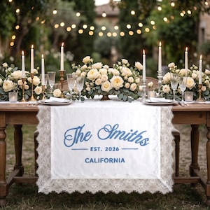 Op de afbeelding: Een prachtig gedekte tafel voor een bruiloft of speciaal evenement. De tafel heeft een witte kanten loper met de tekst "The Smiths EST. 2026 CALIFORNIA" in blauw. Bloemschikkingen, kaarsen en glaswerk maken de elegante setting compleet.