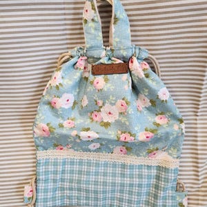 Può includere: Zaino con coulisse floreale con sfondo azzurro e rose rosa. Il manico superiore e il cordino sono beige. La sezione inferiore è un motivo a quadri blu e bianco, con un bordo in pizzo bianco. È attaccata una piccola etichetta in pelle marrone.