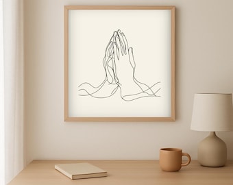 Op de afbeelding: Een ingelijste kunstdruk met een minimalistische lijntekening van handen in gebed. Het zwarte kunstwerk staat tegen een crèmekleurige achtergrond, in een licht houten frame. De print wordt tentoongesteld op een houten tafel.