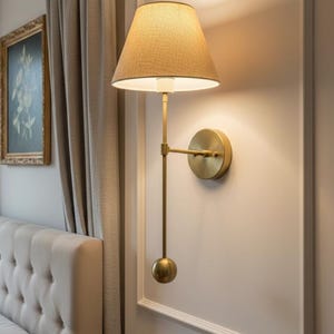 May include: A wall-mounted brass lamp with a beige fabric shade. The lamp has a long, slender arm and a round base. The design is minimalist and elegant, suitable for a bedroom or living room.
