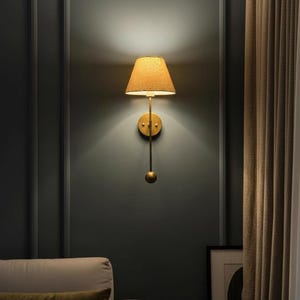 May include: A wall-mounted lamp with a gold finish and a tan lampshade. The lamp has a long, slender arm with a decorative sphere at the bottom. The lampshade is cone-shaped and emits a warm glow. The lamp is mounted on a dark gray wall.