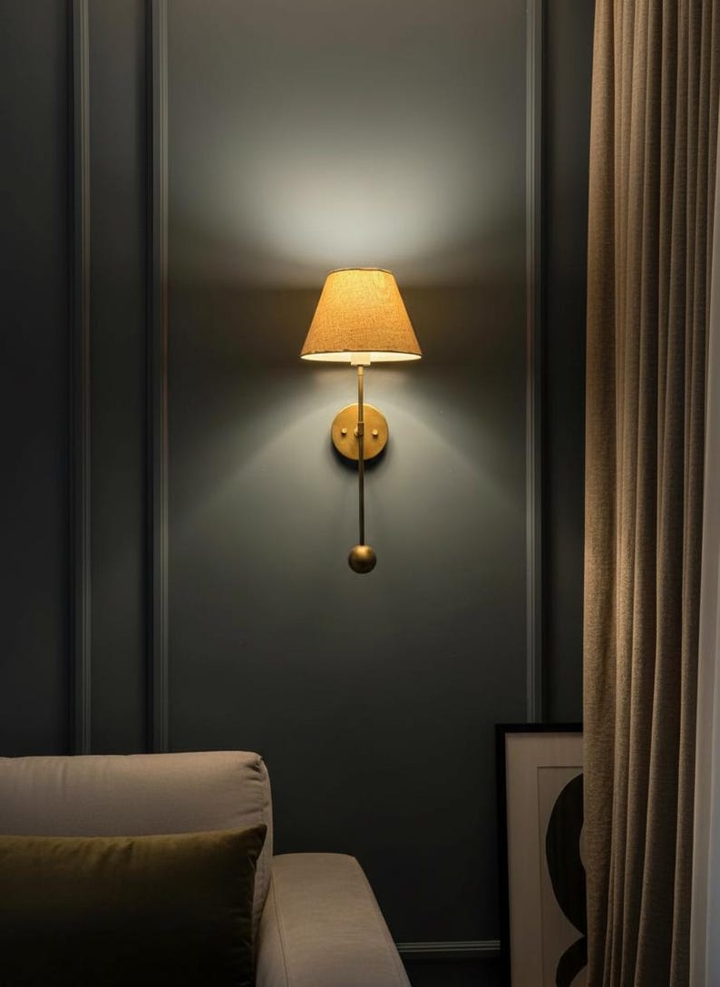 May include: A wall-mounted lamp with a gold finish and a tan lampshade. The lamp has a long, slender arm with a decorative sphere at the bottom. The lampshade is cone-shaped and emits a warm glow. The lamp is mounted on a dark gray wall.