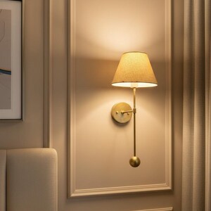 May include: A wall-mounted brass lamp with a beige lampshade. The lamp has a long, slender arm and a round base. The wall behind the lamp is painted a light beige color, and the lamp is positioned in a decorative panel.