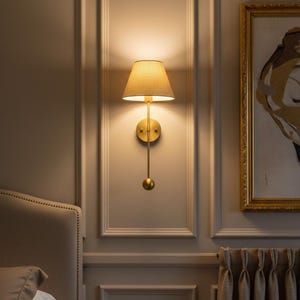 May include: A wall-mounted brass sconce with a beige lampshade. The sconce has a long, slender arm ending in a decorative sphere. The light fixture is mounted on a neutral-colored wall with decorative molding.