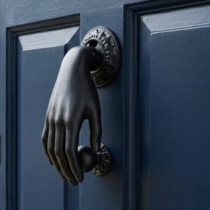 May include: A navy blue door featuring a black hand-shaped door knocker. The door knocker is a detailed hand holding a ball. The door has a keyhole and a black handle. The background shows a stone building and potted flowers.