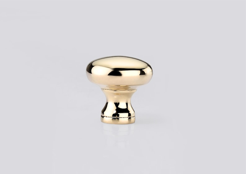 May include: A polished gold-tone cabinet knob featuring a smooth, rounded mushroom-shaped head and a tapered, flared base. The metallic surface is highly reflective, showing bright highlights and deep shadows across its curved, circular form.