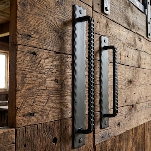 May include: Two black metal door handles with a textured, hammered finish mounted on a rustic wooden door. The handles feature a long, flat backplate with scalloped edges and a cylindrical, knurled grip section attached to the door surface.