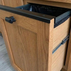 May include: A pull-out kitchen cabinet drawer featuring a light brown oak wood finish and a black T-shaped handle. The drawer is partially extended, revealing a black plastic bin liner inside a dark gray frame with metal sliding rails on the side.