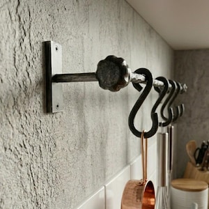 Puede incluir: Un estante para utensilios de cocina de hierro forjado montado en una pared gris texturizada. El estante presenta una pieza final decorativa y varios ganchos en forma de S. Una taza medidora de cobre cuelga de uno de los ganchos.