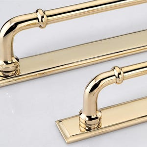 May include: A set of polished gold-toned metal cabinet pulls, each featuring a long, flat rectangular backplate and a rounded, tubular handle attached with two vertical supports. The hardware has a smooth, reflective finish and clean lines.