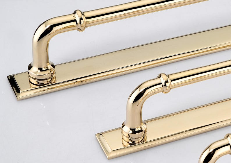 May include: A set of polished gold-toned metal cabinet pulls, each featuring a long, flat rectangular backplate and a rounded, tubular handle attached with two vertical supports. The hardware has a smooth, reflective finish and clean lines.