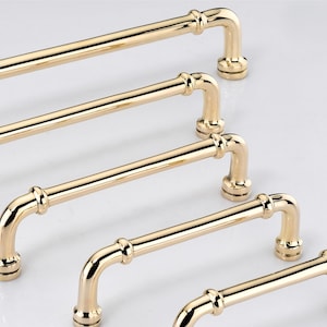 May include: A set of five polished gold-tone metal cabinet pulls arranged in a diagonal row. Each handle features a cylindrical bar with rounded ends, two decorative rings near the mounting points, and circular bases for attachment to furniture.