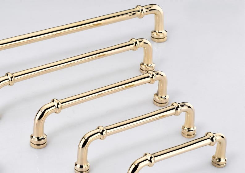 May include: A set of five polished gold-tone metal cabinet pulls arranged in a diagonal row. Each handle features a cylindrical bar with rounded ends, two decorative rings near the mounting points, and circular bases for attachment to furniture.