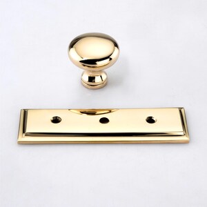 Puede incluir: Un pomo de mueble de metal en tono dorado pulido con una cabeza lisa y redondeada en forma de hongo y una placa rectangular a juego que presenta tres orificios para tornillos. Los herrajes tienen un acabado reflectante de alto brillo.