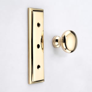 May include: A polished brass door knob and a matching rectangular backplate with three screw holes. The hardware features a reflective, smooth metallic finish and a classic rounded shape, presented as separate components on a plain white background.