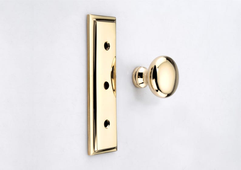 May include: A polished brass door knob and a matching rectangular backplate with three screw holes. The hardware features a reflective, smooth metallic finish and a classic rounded shape, presented as separate components on a plain white background.