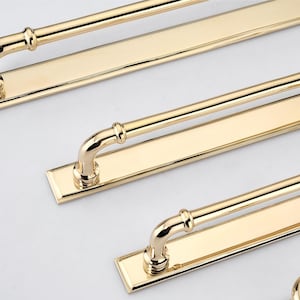 May include: A set of polished gold-tone metal cabinet pulls, each featuring a long rectangular backplate and a rounded handle bar attached with two curved supports. The hardware has a reflective, smooth finish and a clean, linear design.