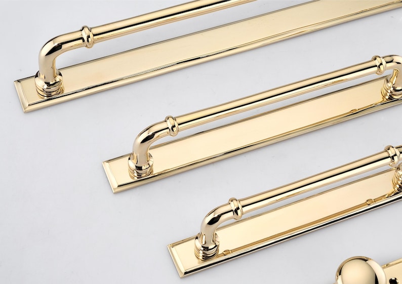 May include: A set of polished gold-tone metal cabinet pulls, each featuring a long rectangular backplate and a rounded handle bar attached with two curved supports. The hardware has a reflective, smooth finish and a clean, linear design.