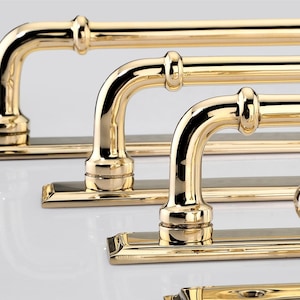 May include: A series of polished brass door handles and a matching round knob, each mounted on a rectangular backplate. The hardware features a reflective, metallic gold finish with curved, tubular grips and decorative collar details.