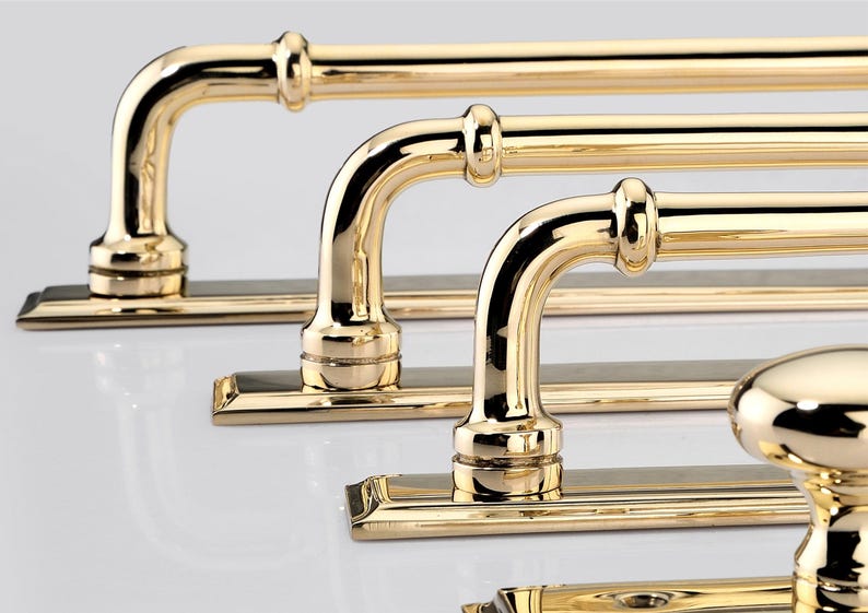 May include: A series of polished brass door handles and a matching round knob, each mounted on a rectangular backplate. The hardware features a reflective, metallic gold finish with curved, tubular grips and decorative collar details.